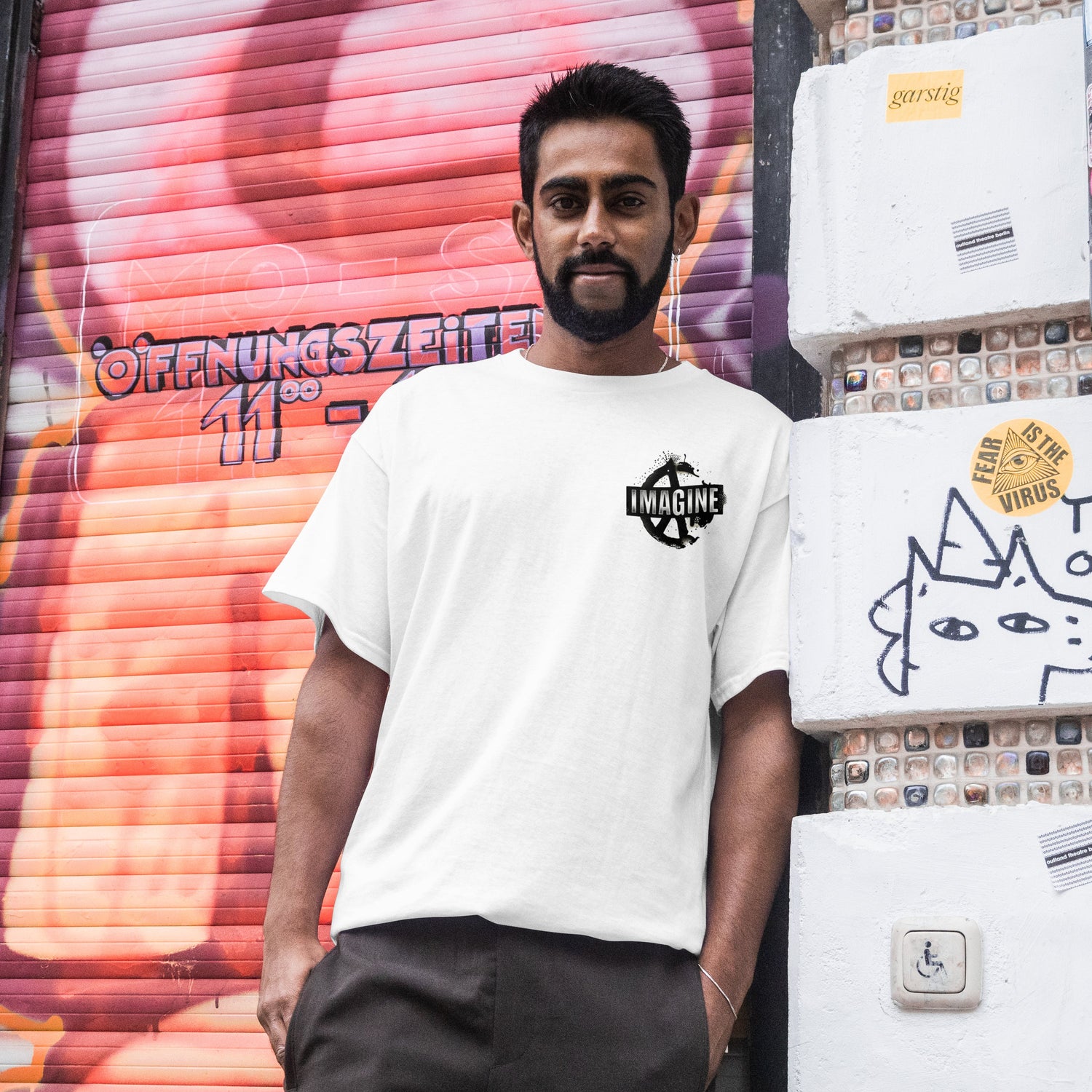 Ein Mann steht lässig an eine Wand gelehnt und trägt ein weißes T-Shirt mit einem "Imagine Peace" Motiv
