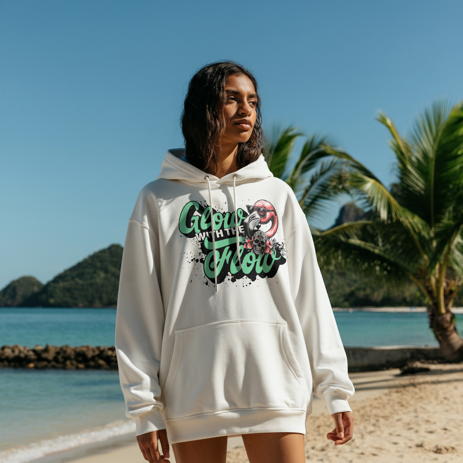 Eine Frau am Strand trägt einen weißen Hoodie mit einem Flamingo Print und den Worten "Glow with the Flow"