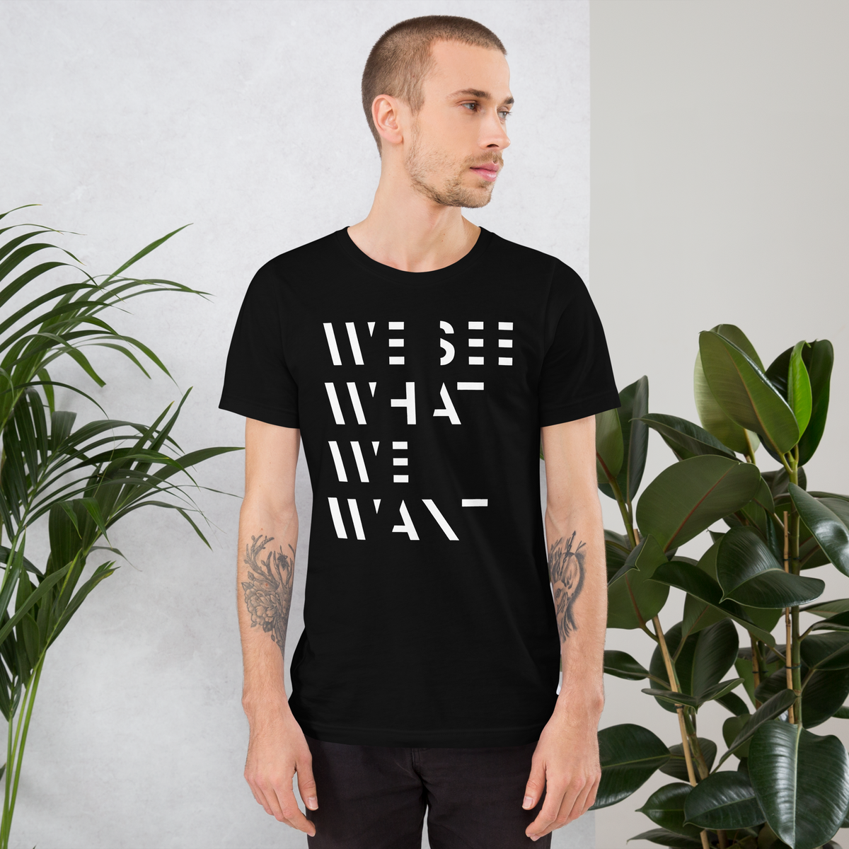 Mann trägt Schwarzes Unisex T-Shirt mit We See What We Want Print bei dem die Buchstaben teilweise ausgeschnitten sind